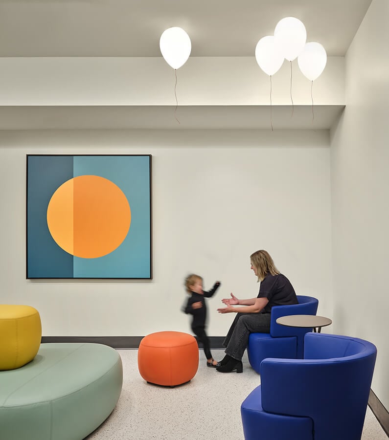 Modern room with colorful seating, abstract art, and balloon-shaped lights. Child and adult interacting.