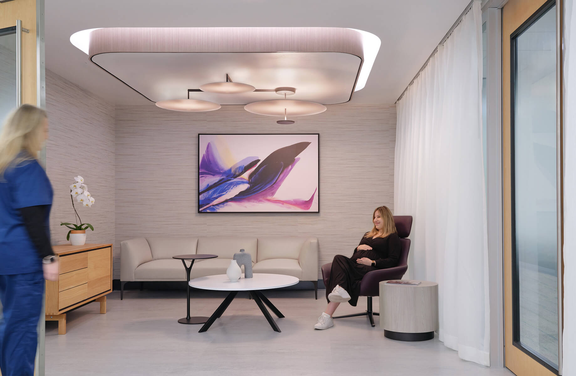 Pregnant woman sits in modern waiting room with abstract art, diffused overhead light, and comfortable seating.