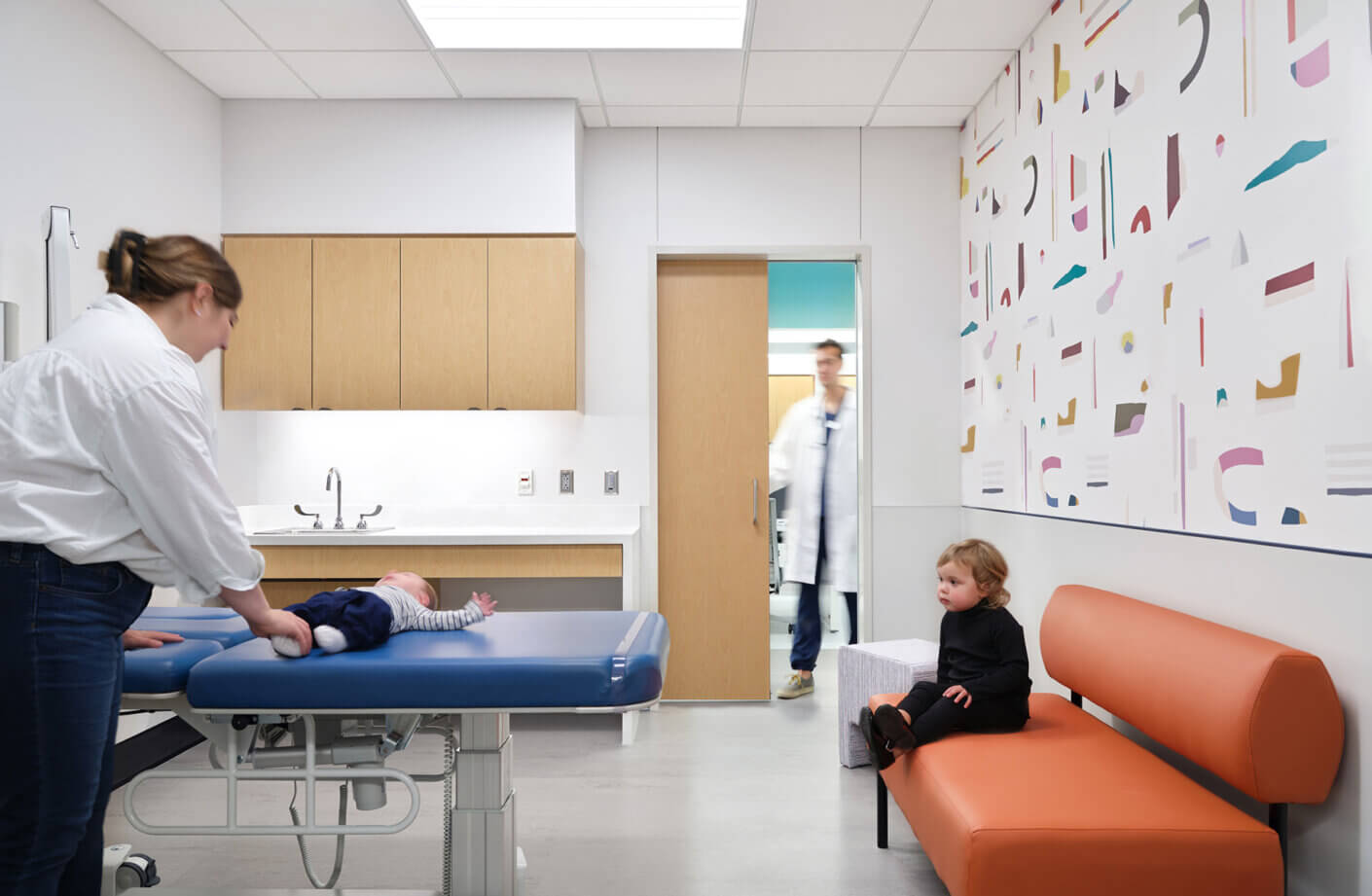 Patient room with abstract wall art, colorful seating, and doctor entering from staff area visible behind.