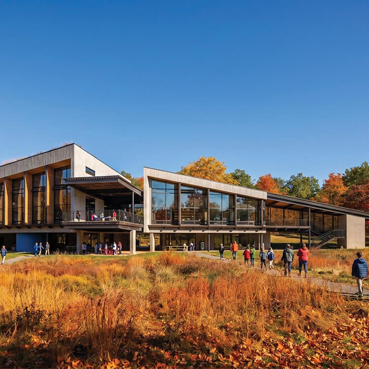 Exterior view of a building with large glass windows, surrounded by colorful autumn foliage and people walking in the foreground.