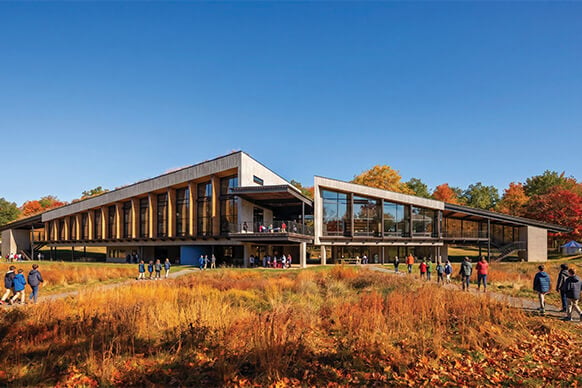 Visitors walk toward a building with large glass windows and wooden accents, surrounded by autumn foliage and clear blue skies.