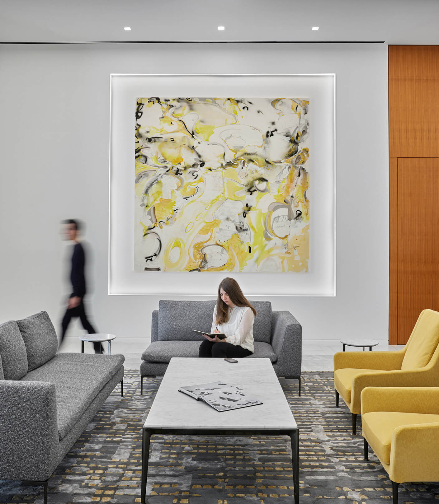 person sitting in chair and person walking past large art piece on back wall