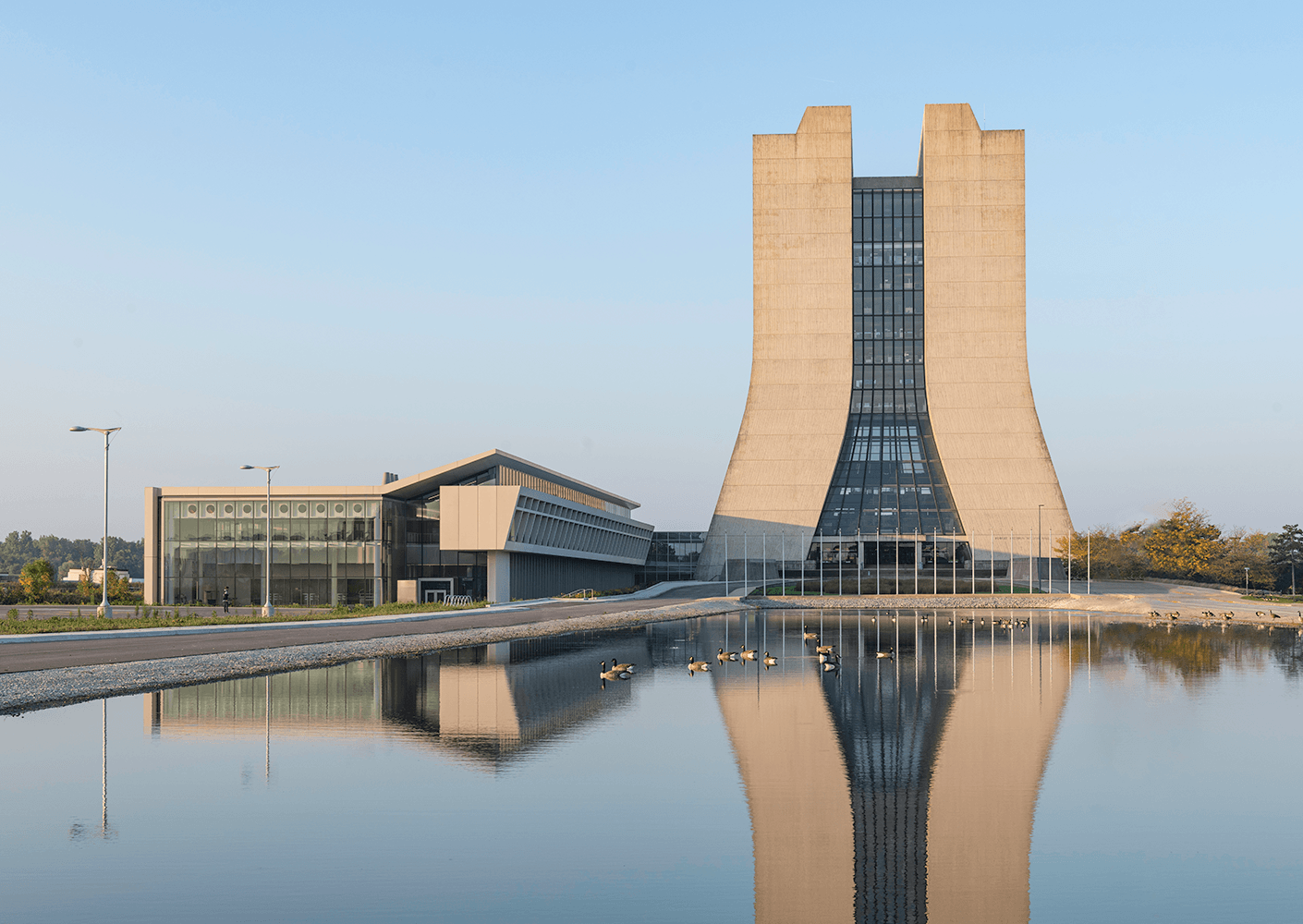 Concrete building with large glass sections, reflecting in a calm body of water. Surrounding area features greenery and light poles.