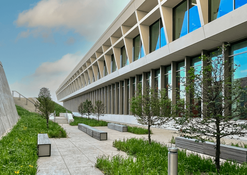 Exterior view of a building featuring large windows, geometric design elements, landscaped seating area, and walking pathways surrounded by greenery.
