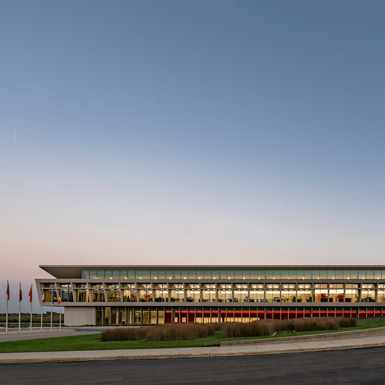 A contemporary building with large windows, landscaping, and flags displayed in front, under a clear sky at dusk.