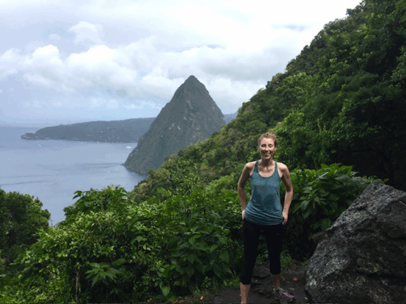 Woman standing on a mountain surrounded by trees with mountains and water in the distance.