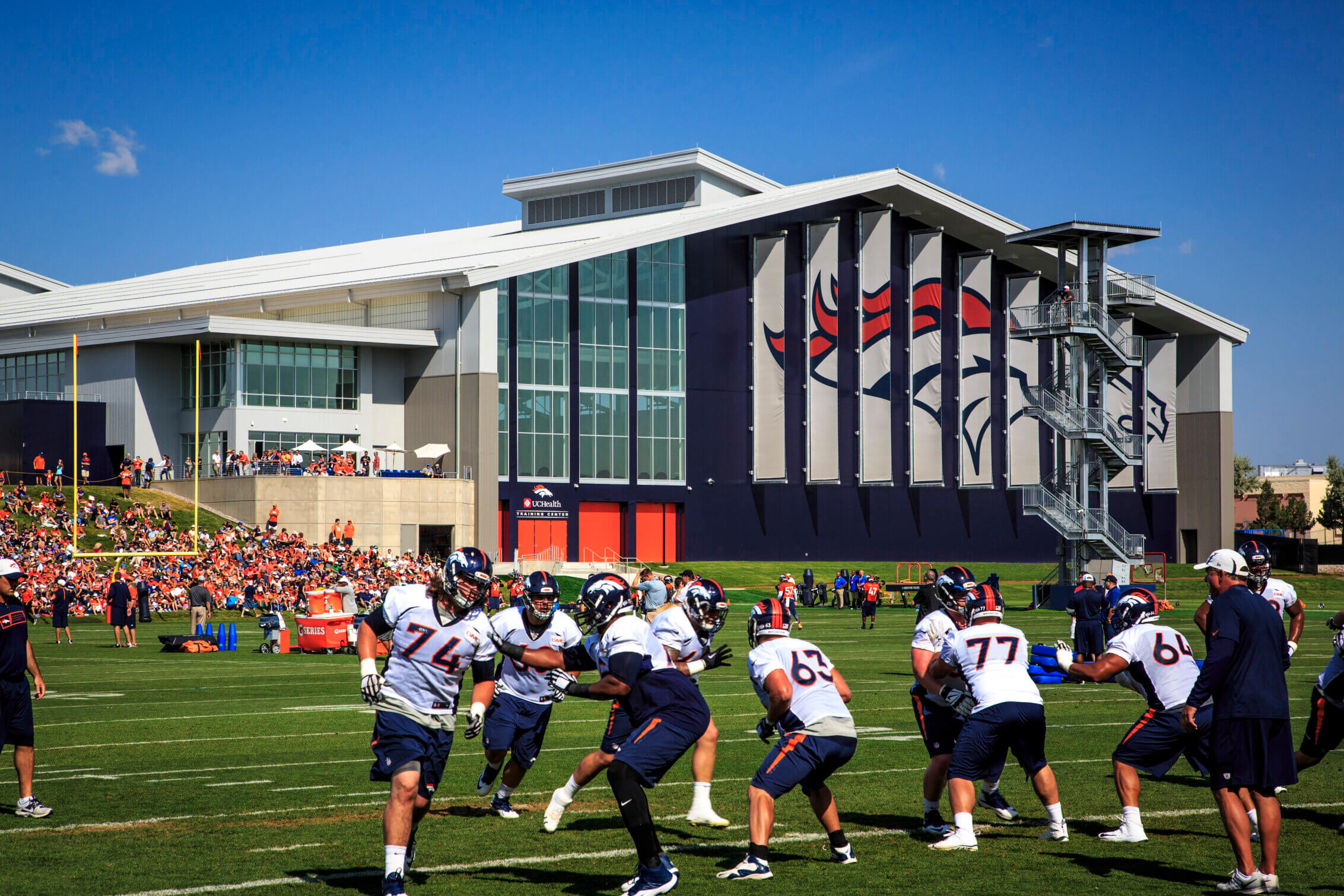 Football players in Denver Broncos uniforms practice on a green field in front of a modern training facility with large glass windows and a prominent Broncos logo, while a crowd of spectators watches from the sidelines under a clear blue sky.