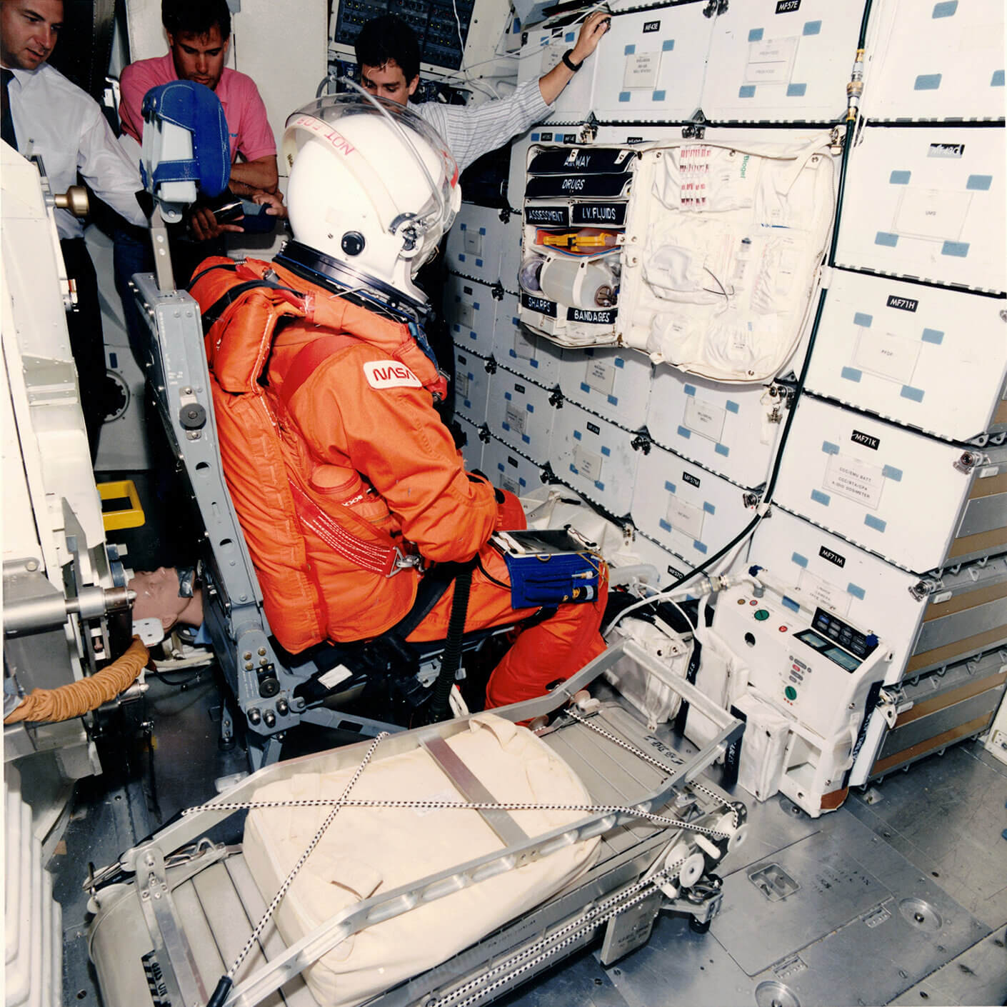 three men with astronaut in space suite inside space station mock up