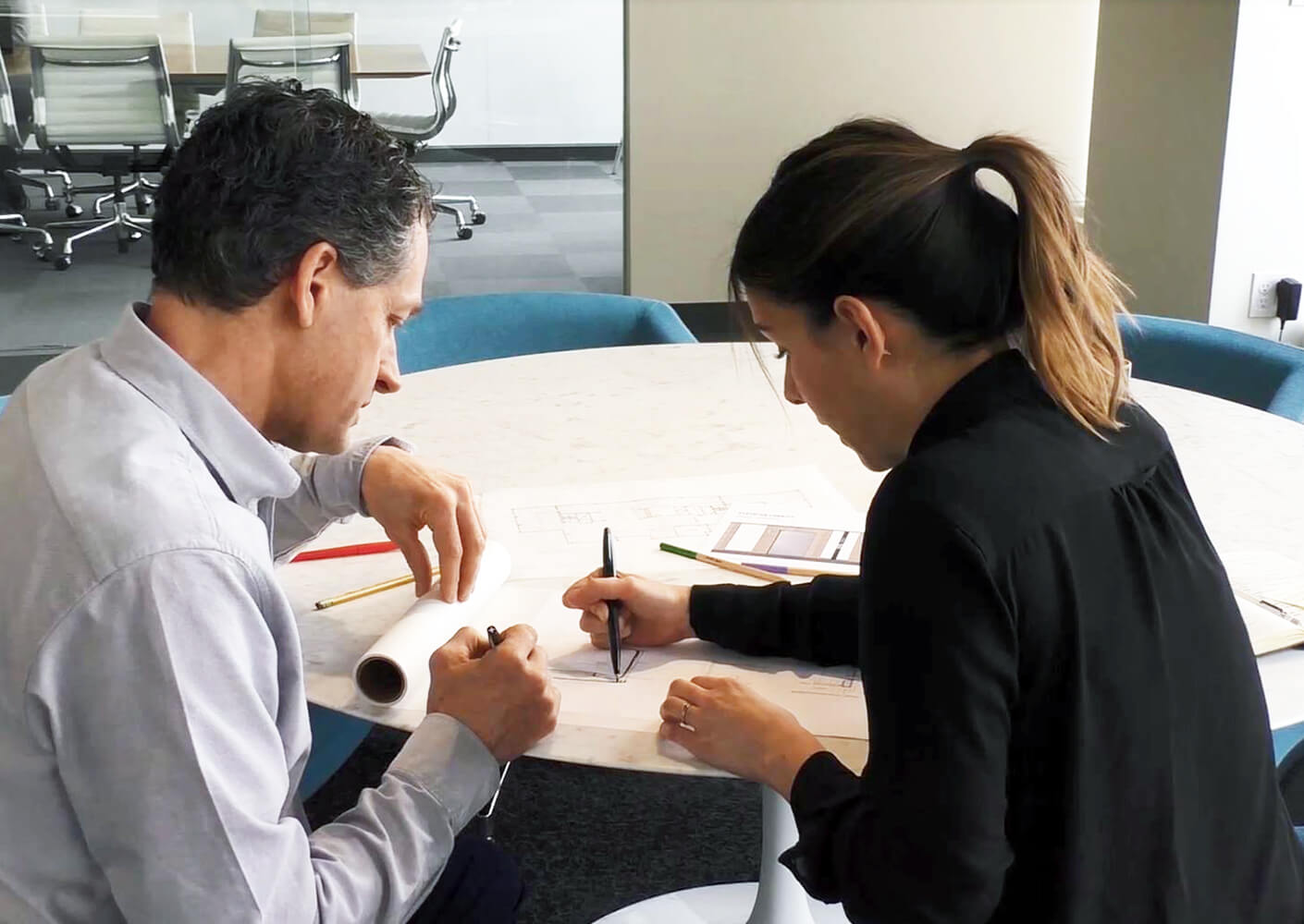 Two architects collaborate on blueprints at a round table in a modern office setting.