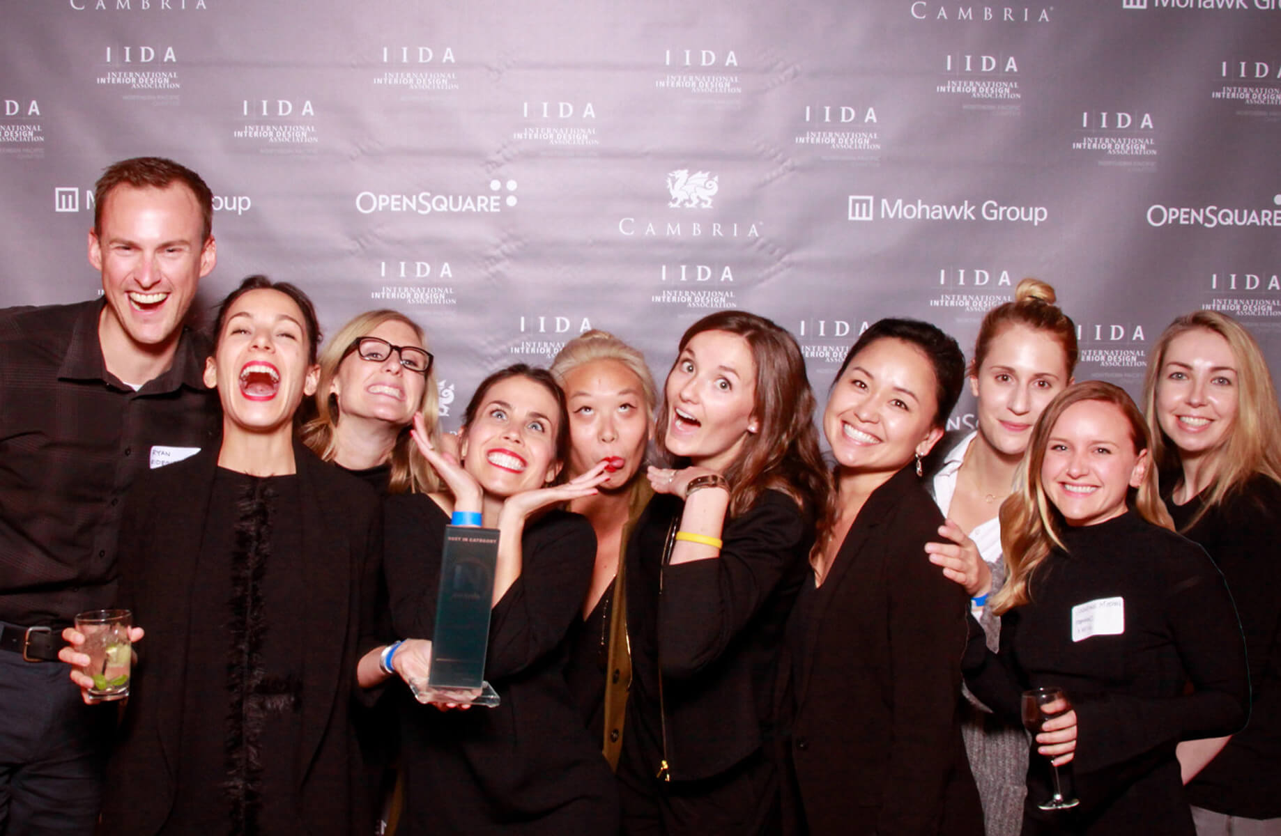 Group celebrating at IIDA event with awards and drinks, joyful expressions against branded background.
