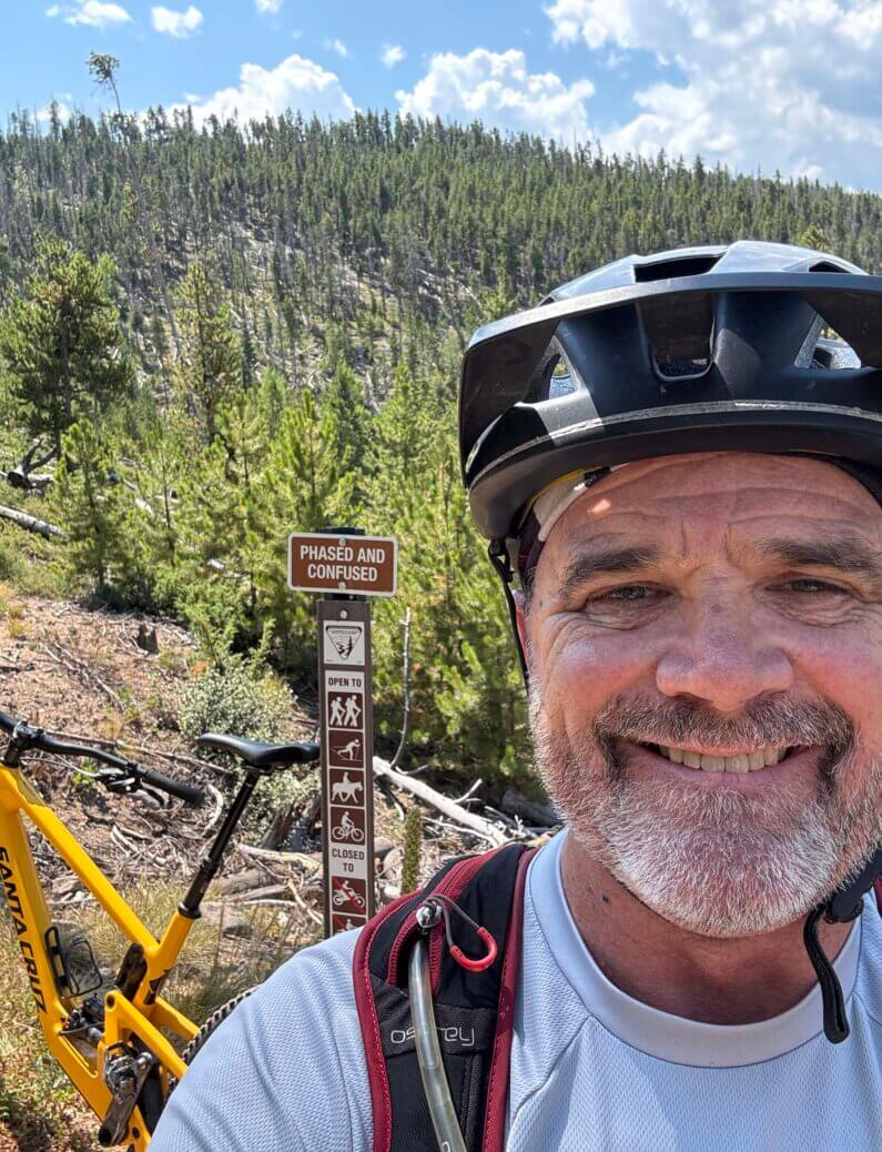 Smiling Steve King wearing a bike helmet and hydration pack takes a selfie on a forested mountain trail, with a yellow Santa Cruz mountain bike and a trail sign reading 'Phased and Confused' in the background.