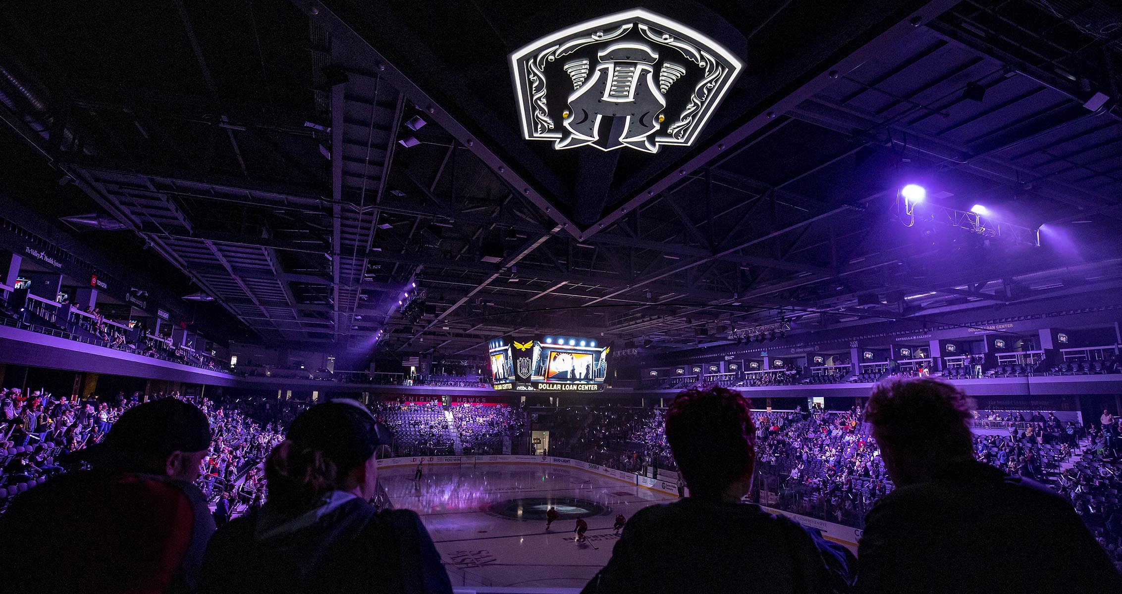 Fans watch from the stands as a hockey game is about to begin at the arena, with purple lighting, a large center-hung scoreboard, and the team logo illuminated on the ceiling.