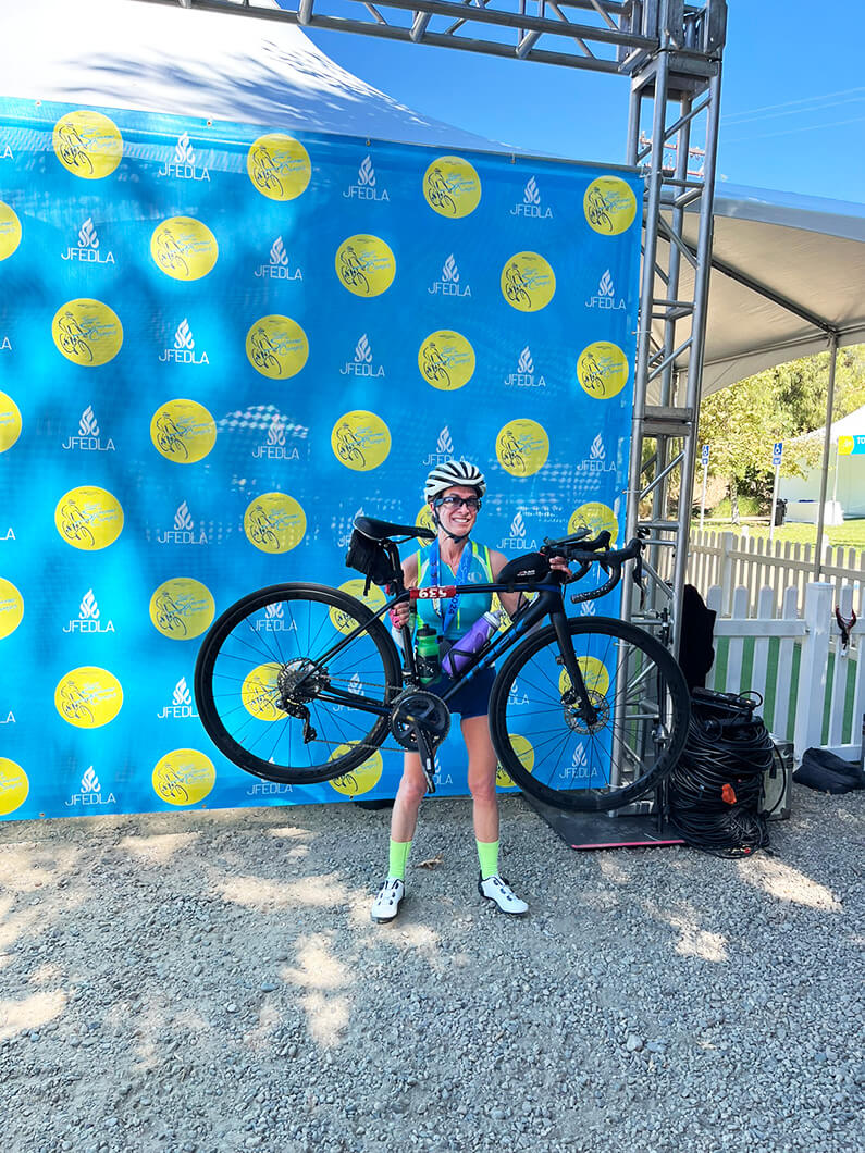 Carolyn posing with bike in front of JFEDLA backdrop at outdoor event.