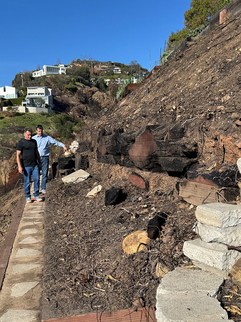 Two men examining a hillside with burnt vegetation, with houses visible in the background on a sunny day.