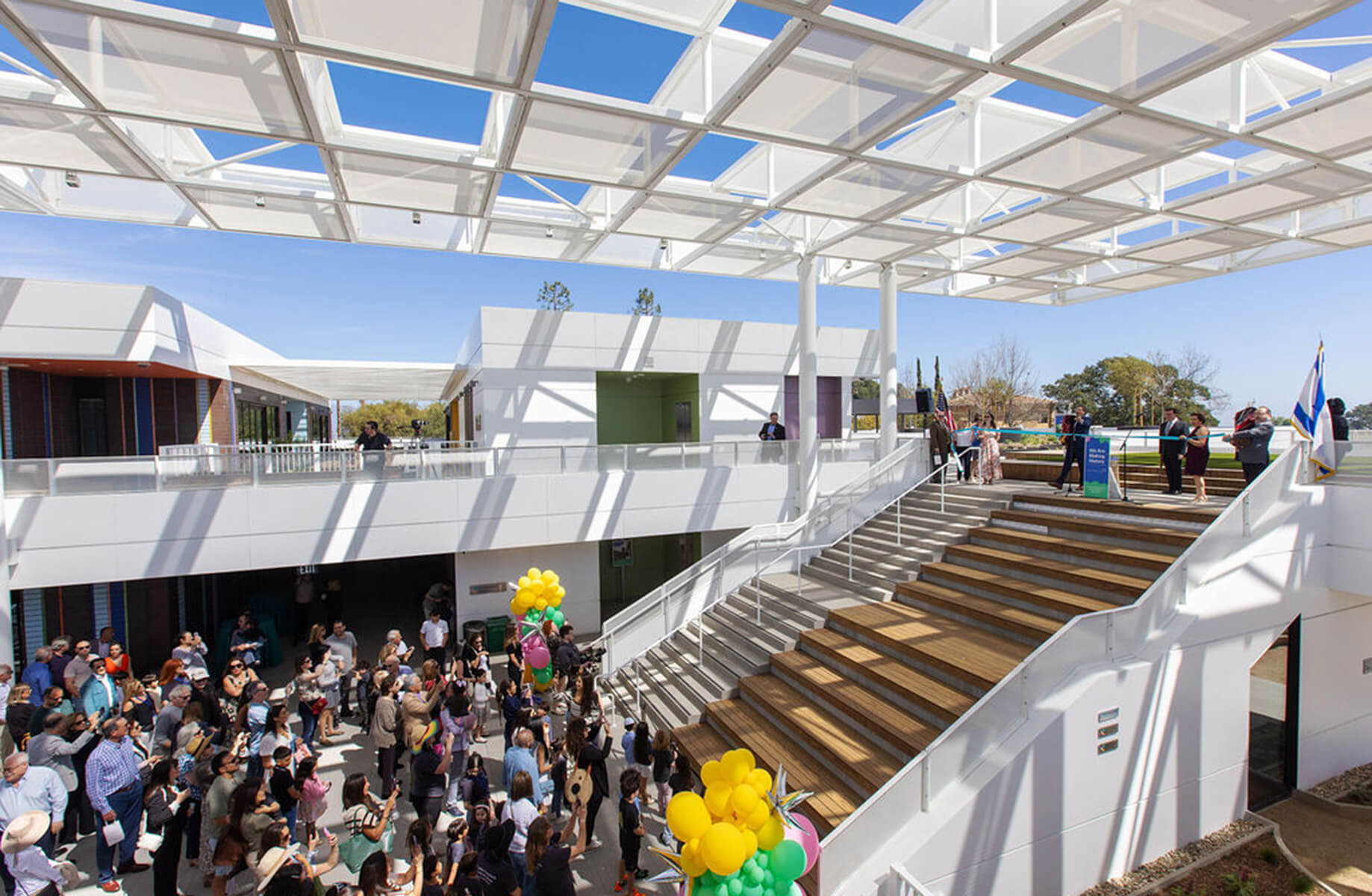 Modern open-air atrium with a crowd attending a ribbon cutting event, featuring balloons, stairs, and a geometric ceiling design.