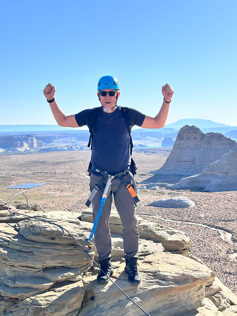 Trevor celebrating on a rocky mountain summit, wearing climbing gear and a blue helmet, with a scenic landscape view.