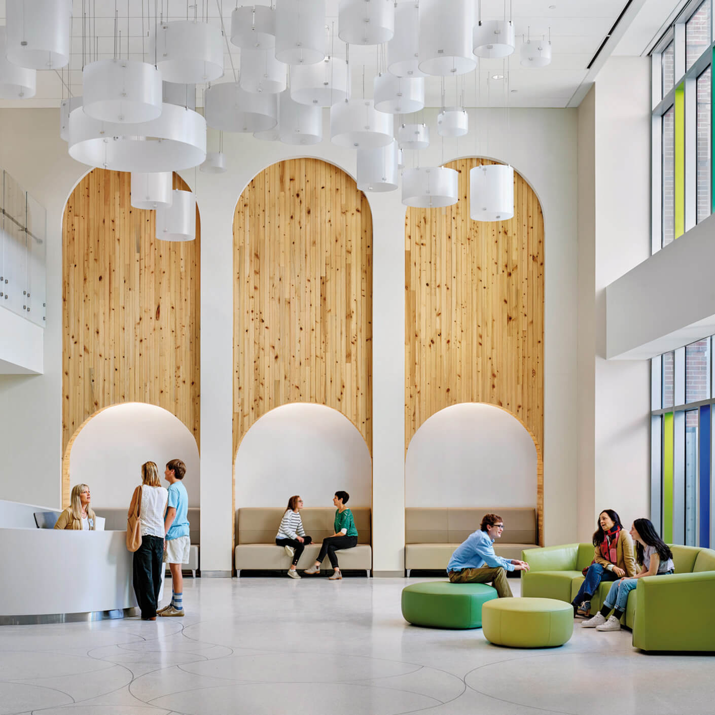 Modern lobby with wood accents, people conversing and sitting on green furniture, high ceilings with white cylindrical lights.