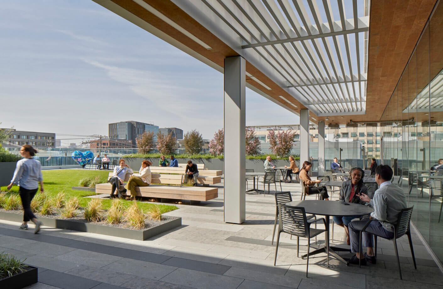 Rooftop terrace with people relaxing, chatting, and working under a modern pergola in an urban setting.