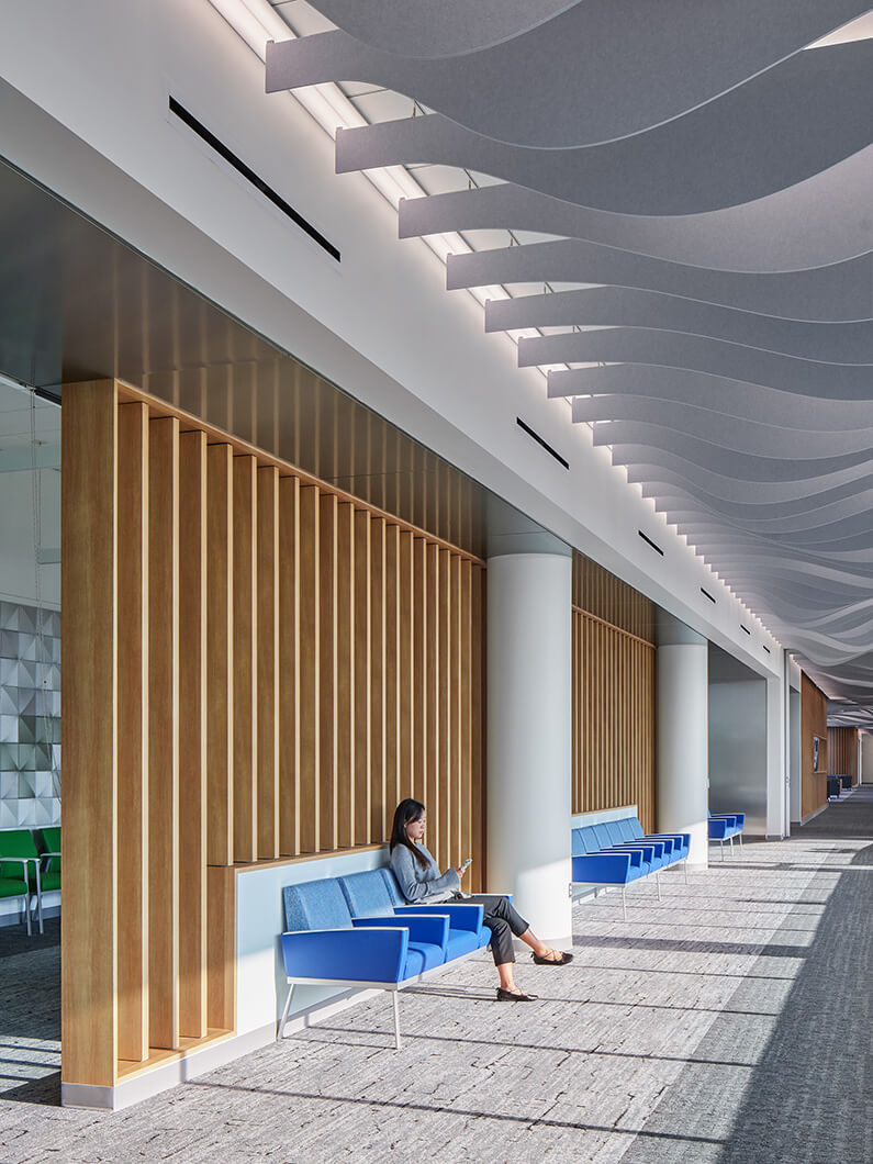 Modern office interior with wooden accents, blue seating, and a person using a phone in a bright hallway.