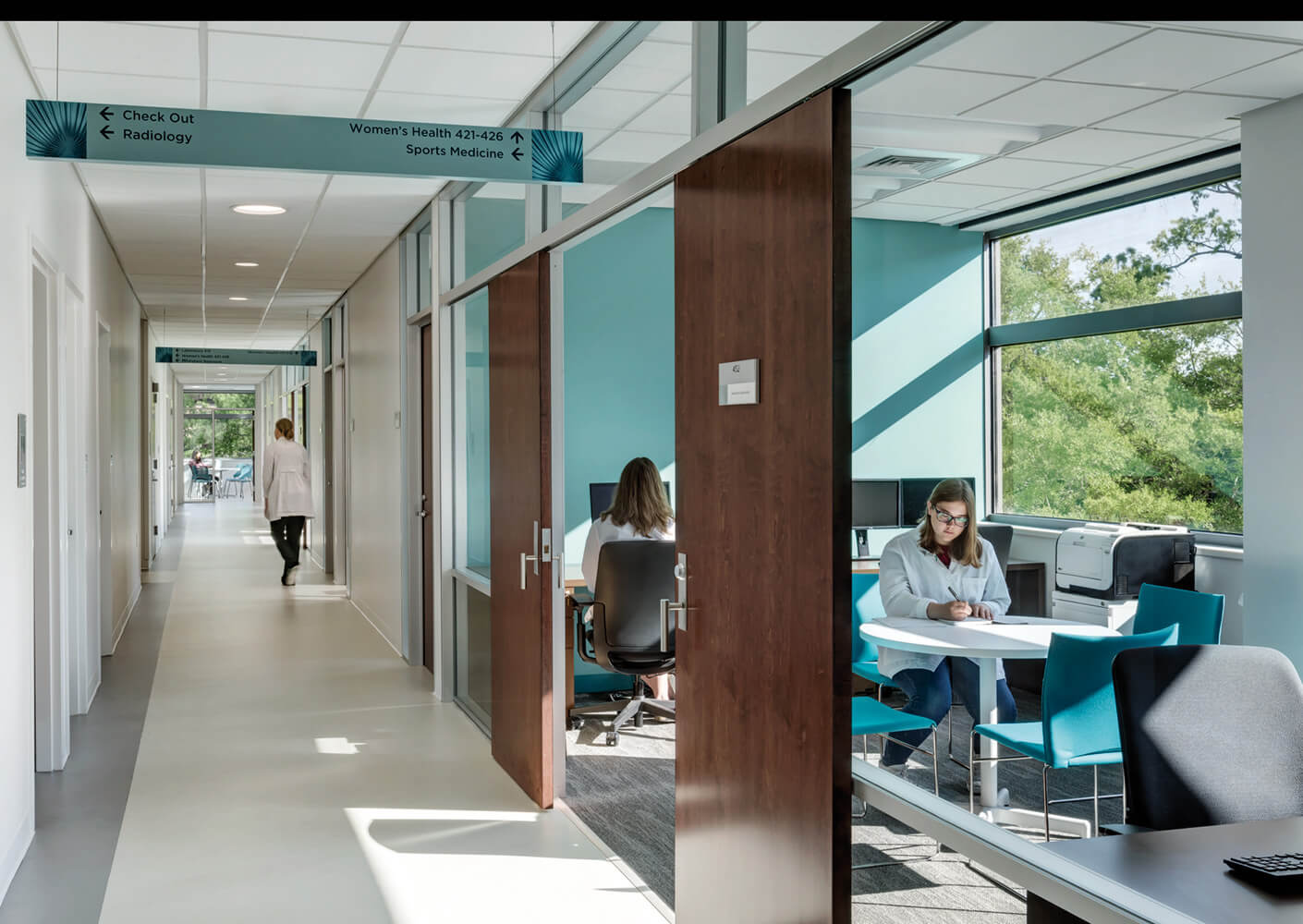 Modern medical office hallway with directional signs, staff working in glass-walled room, natural light from large windows.