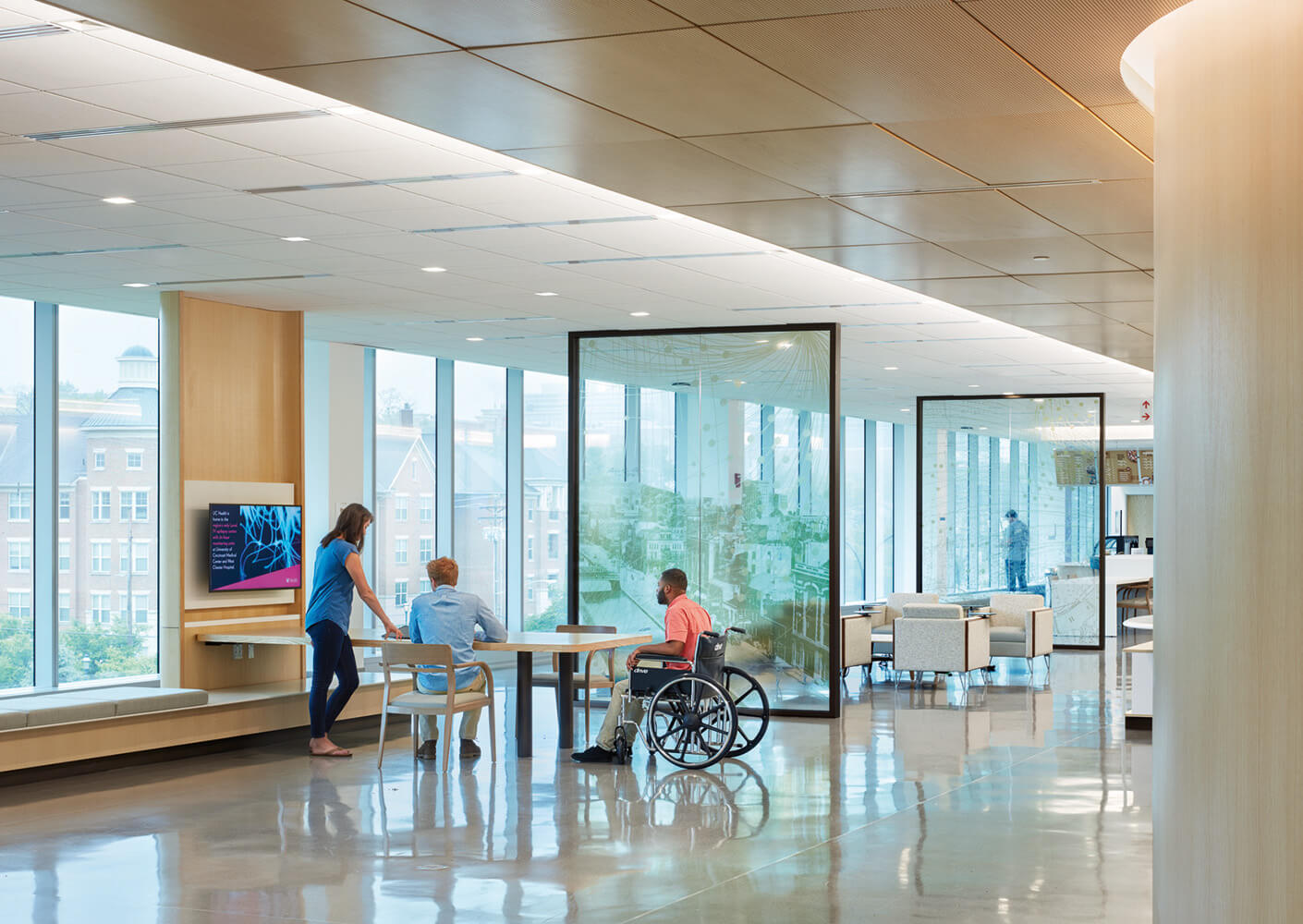 Modern hospital lounge with patients, wheelchair, and staff discussing care. Bright, spacious interior with large windows.