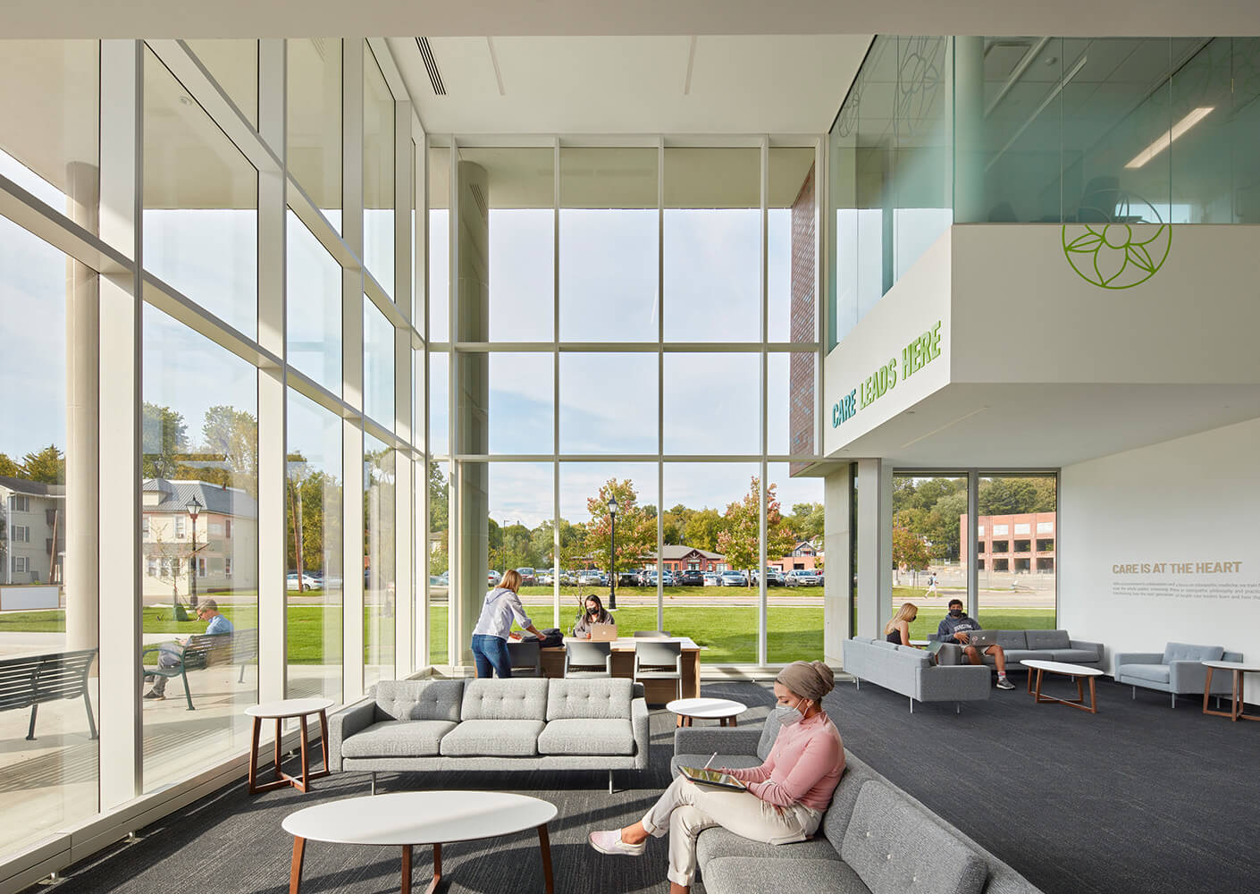 Modern hospital lobby with large windows, people socializing, and Care Leads Here sign on the wall.