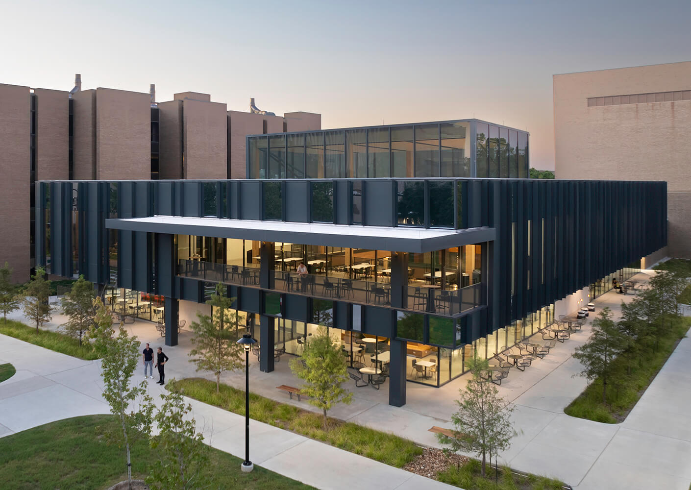 Modern university building with glass facade, surrounded by trees and pathways, viewed at dusk.