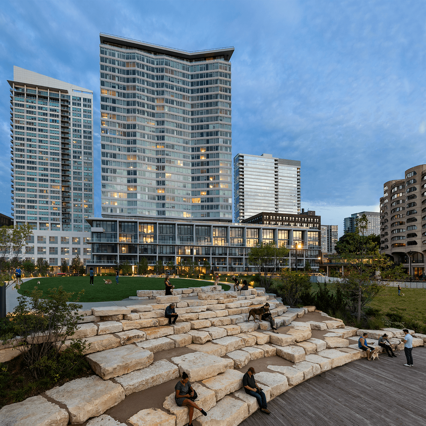 People relax on stone seating in a modern urban park surrounded by tall buildings under a blue sky.