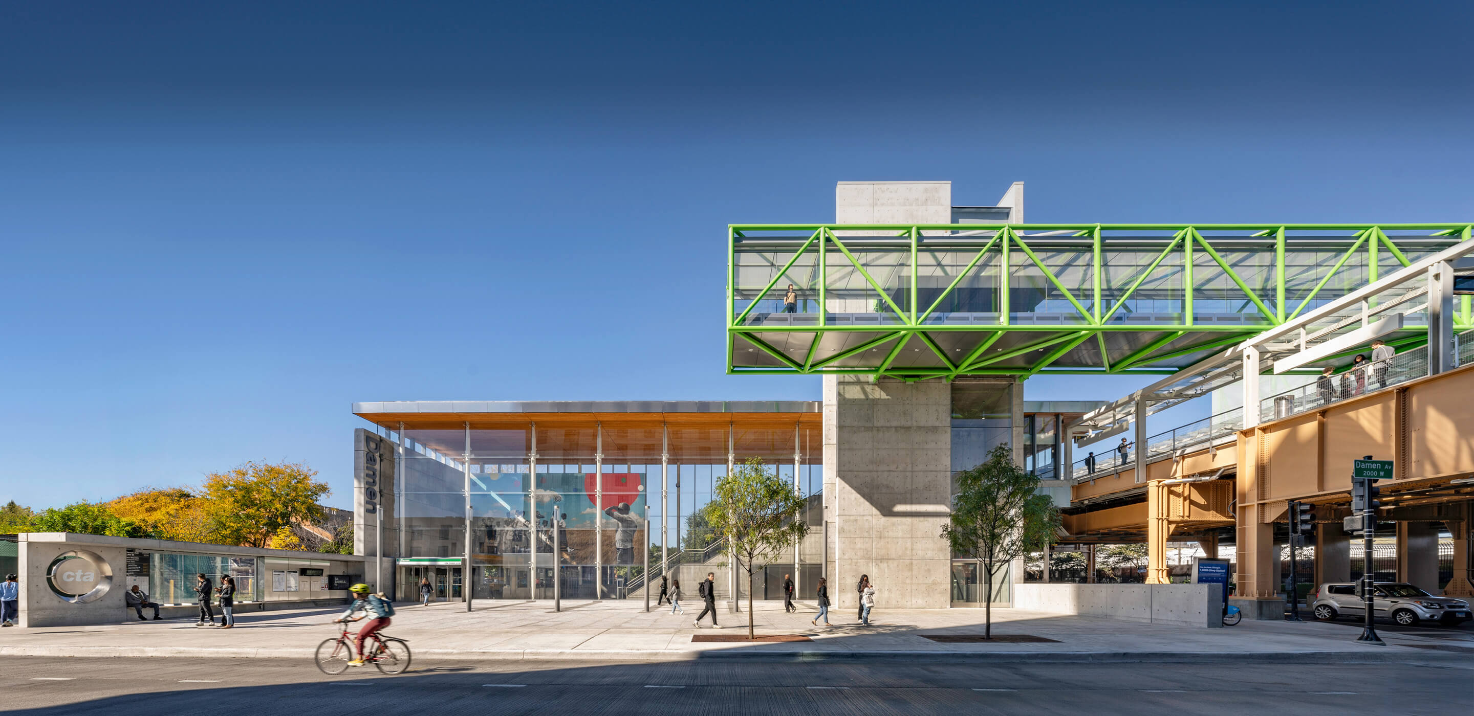 Modern transit station with green walkway, cyclists, and pedestrians under a clear blue sky.
