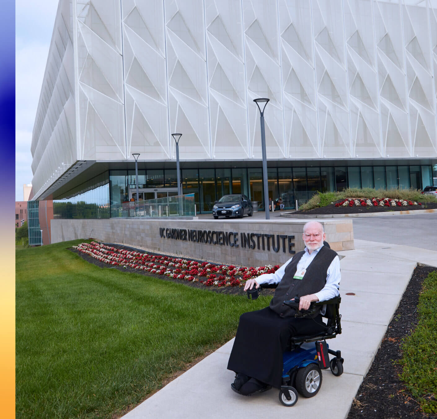 Person in wheelchair outside UC Gardner Neuroscience Institute. Background of modern architecture and flowerbeds.