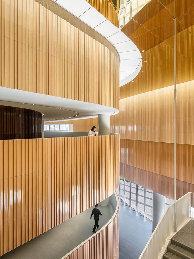 Modern interior with curved wooden panels and people walking on a spiral walkway. Bright natural lighting.