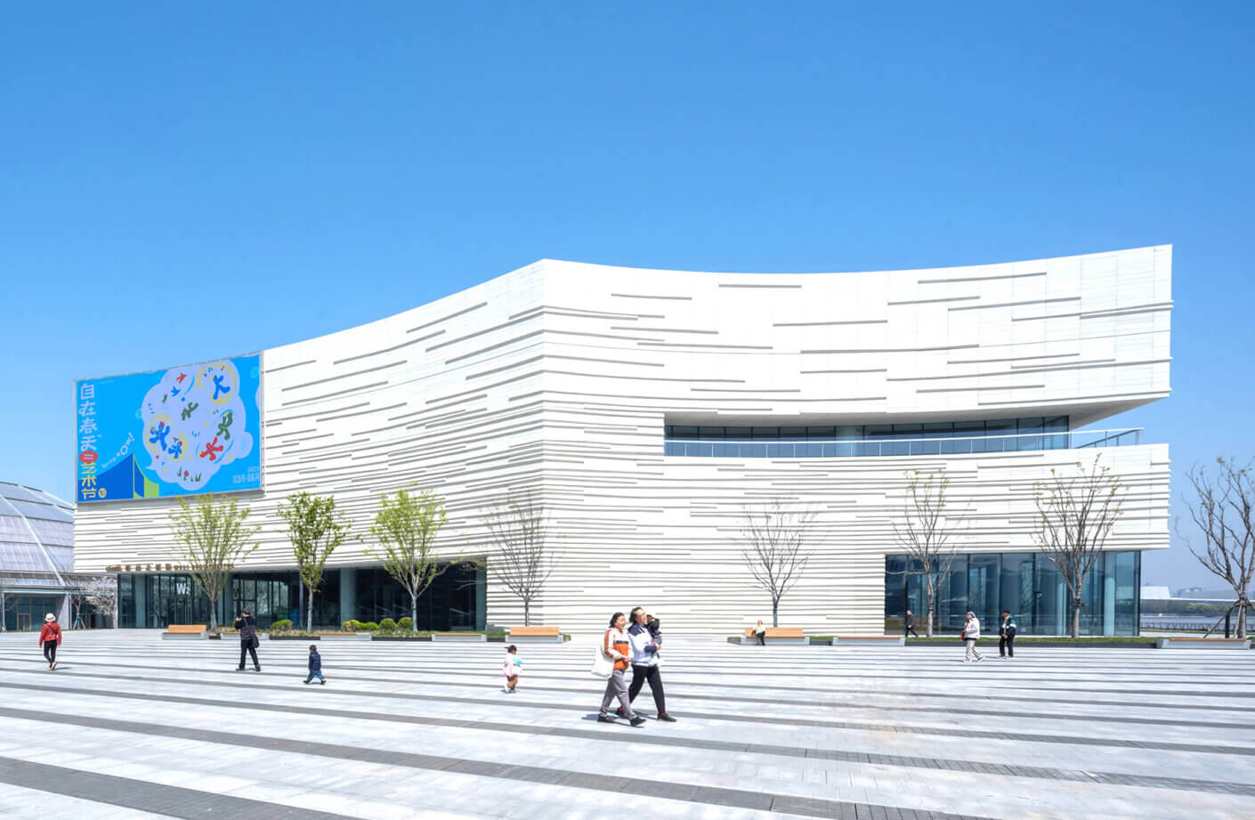 Modern white building with a striped facade under a clear blue sky, people walking in front.