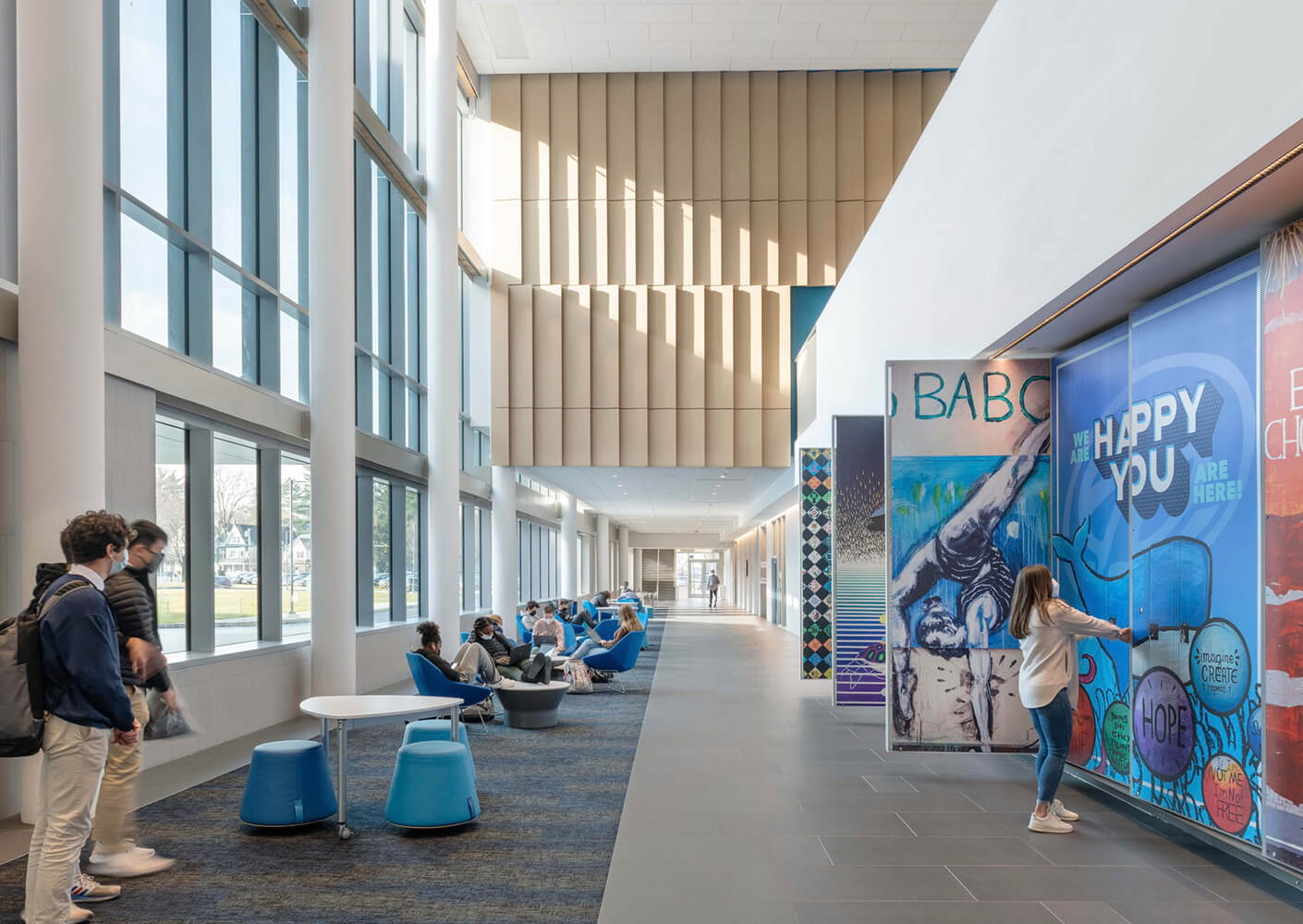 Modern school hallway with students relaxing, colorful wall murals, and large windows for natural light.