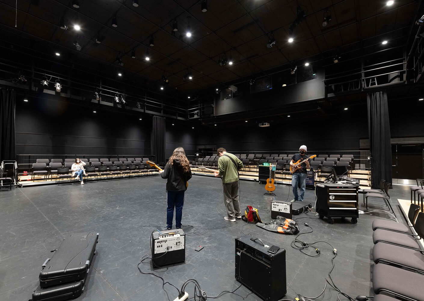 Blackbox theater space with musicians rehearsing with amps and instruments under bright lights.