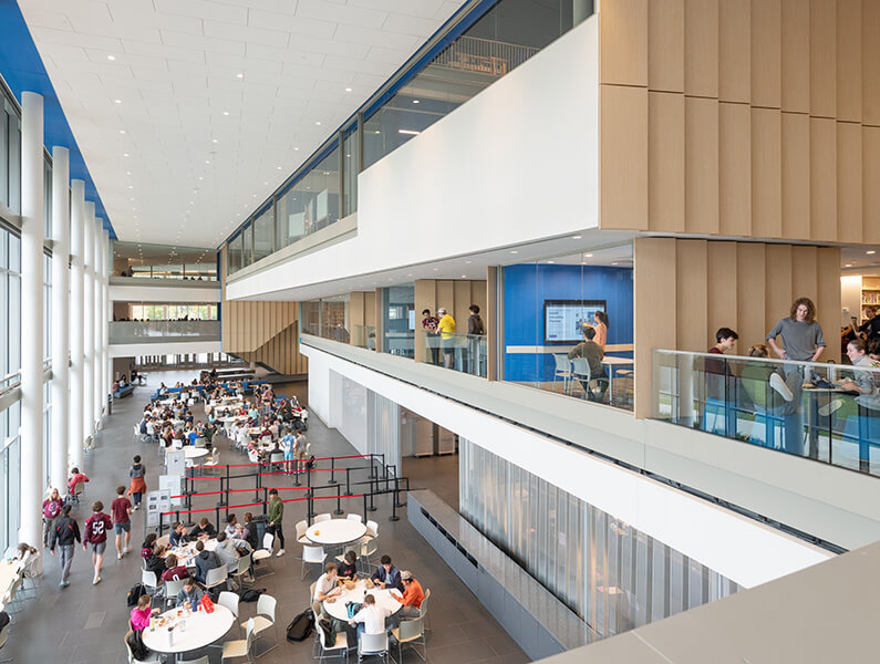 Modern student cafeteria area with tables and seating, large windows, and upper-level walkways with people studying.