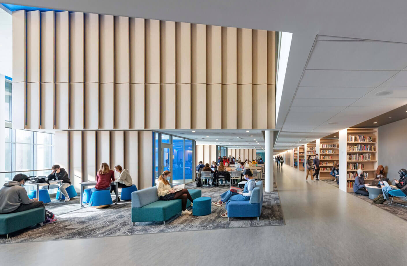 Modern library interior with students studying on blue furniture, large wooden panels, and bookshelves lining the hallway.