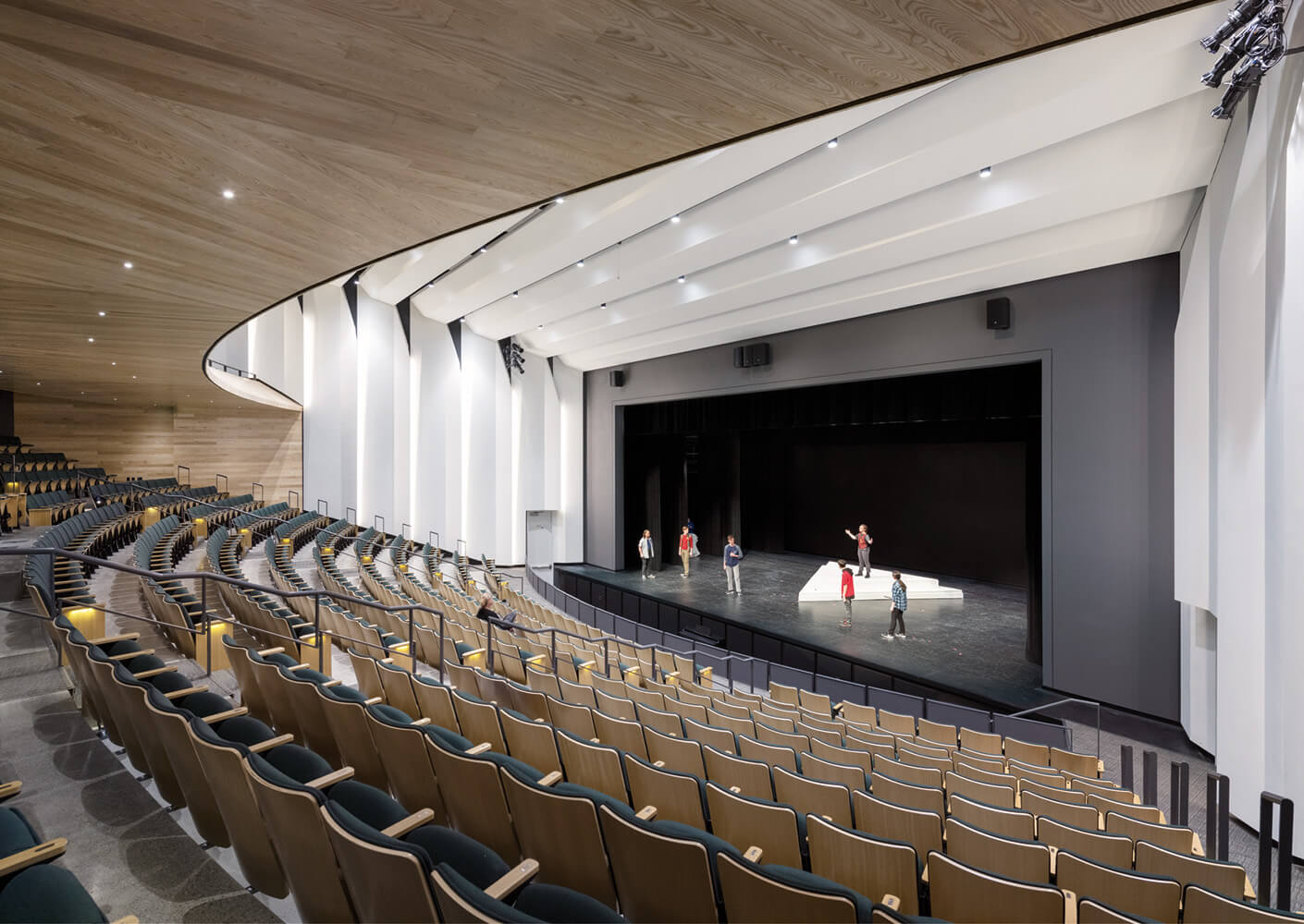 Modern auditorium interior with wooden seats and stage, people rehearsing.