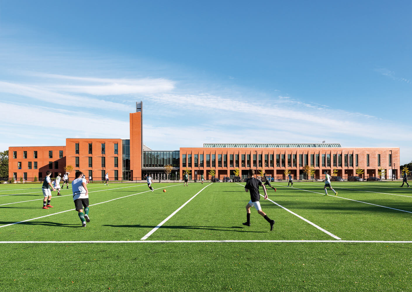 Students playing soccer on a green field in front of a modern red-brick school building under a clear blue sky.