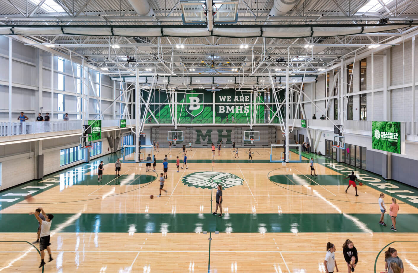 Indoor gym with basketball courts filled with students practicing under high ceilings and bright lighting.