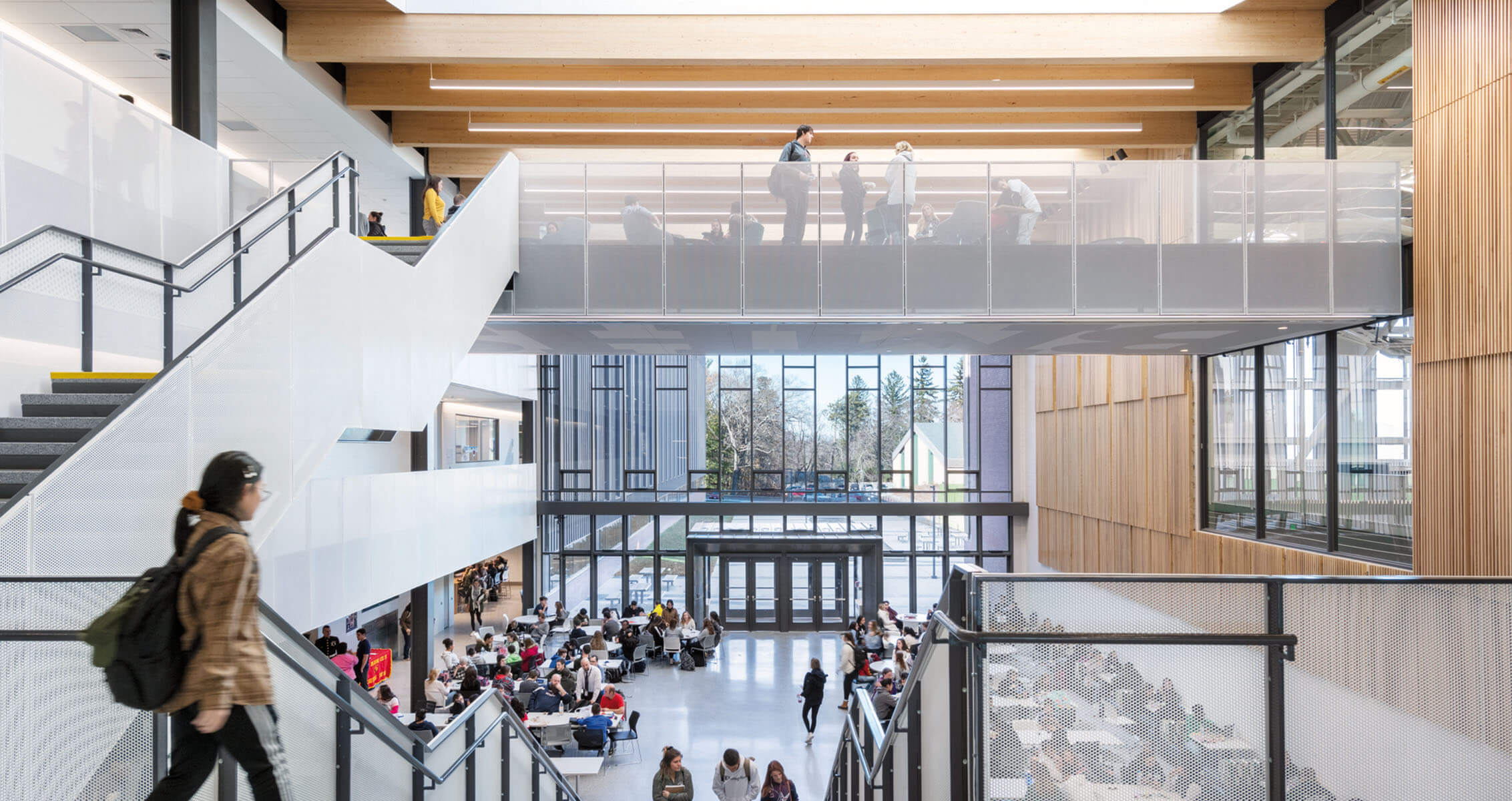 School cafeteria atrium with students walking and studying, featuring large windows and wooden accents.