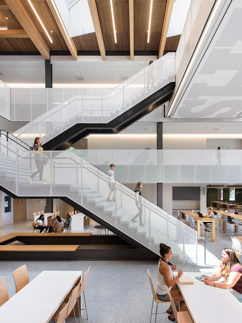 Modern educational space with students on stairs and studying at tables, featuring open layout and natural lighting.