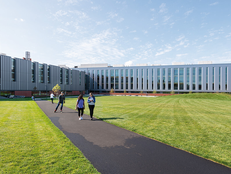 Modern university building with students walking on a path, surrounded by green lawn and clear blue sky.