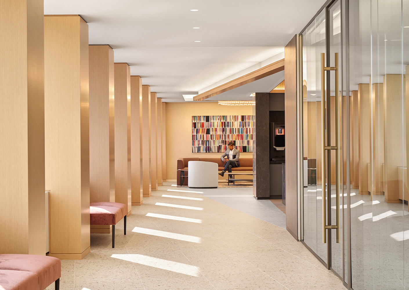 Modern office lobby with wooden columns, pink benches, and a colorful artwork. A person sits on a couch in the background.