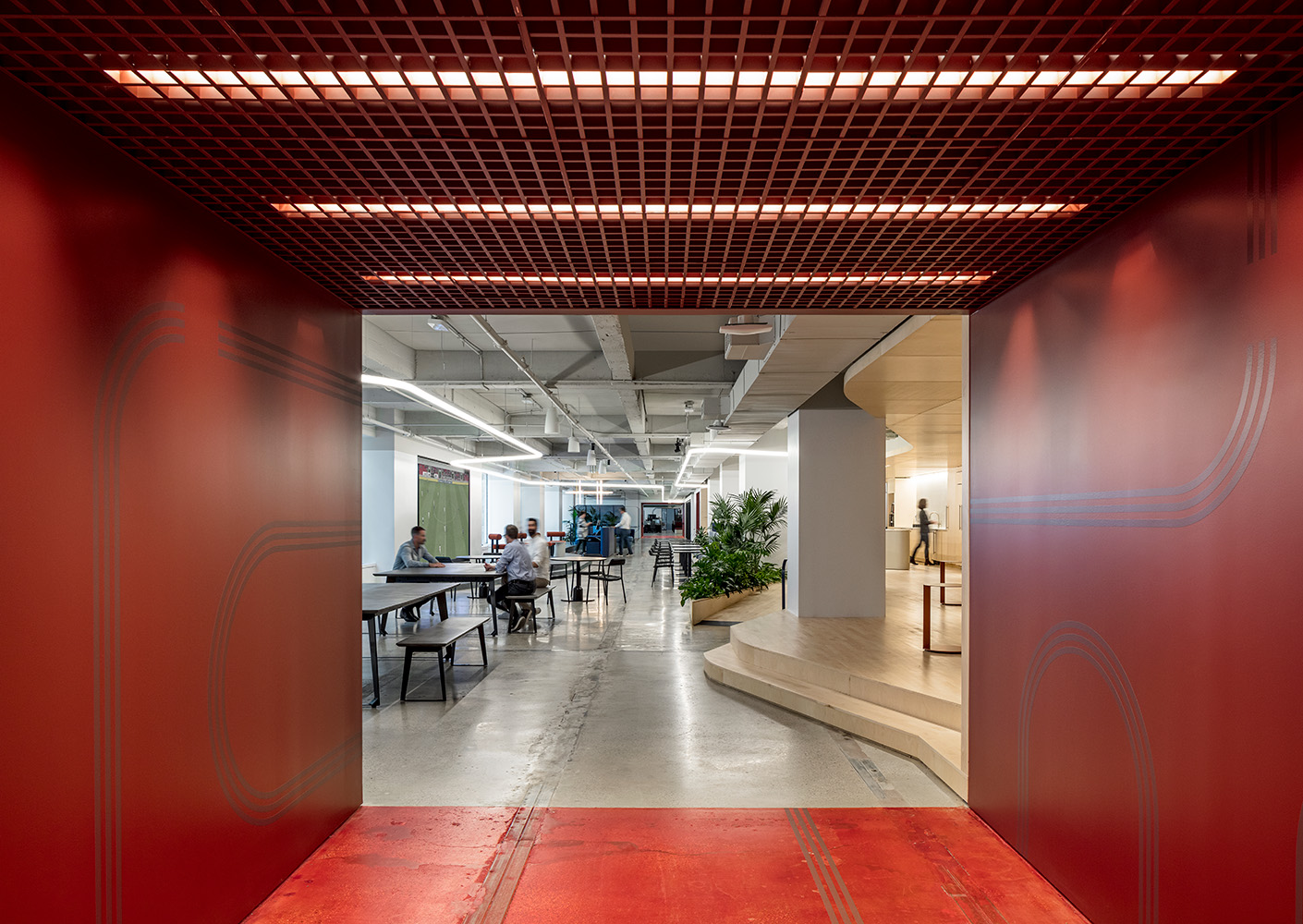 Modern office interior with red entrance, communal tables, plants, and people working creatively.