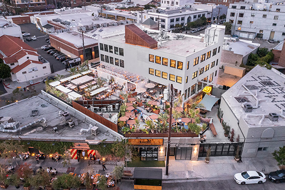 Aerial view of a bustling urban rooftop restaurant with outdoor seating and surrounding cityscape at dusk.