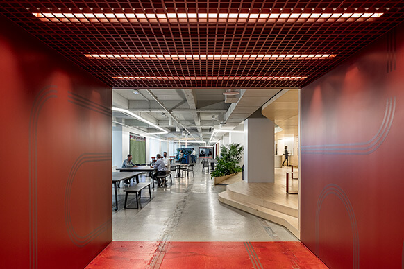 Modern office space with people working at tables, featuring red walls, open ceiling, and indoor plants.