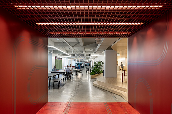 Modern office space with people working at tables, featuring red walls, open ceiling, and indoor plants.