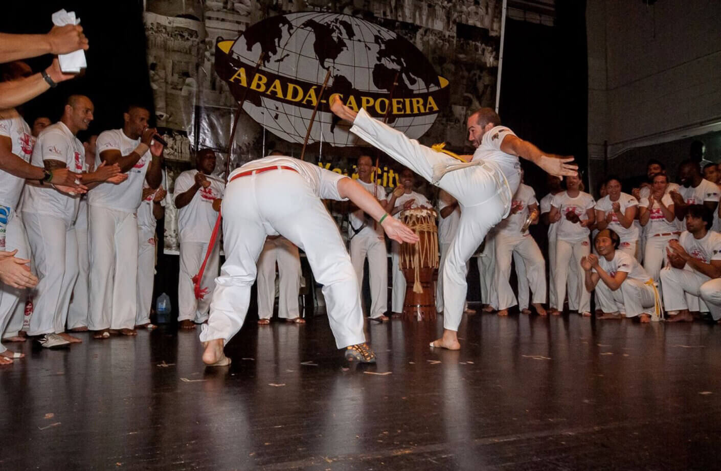 Two capoeira practitioners in action at an Abadá-Capoeira event, audience clapping and cheering.