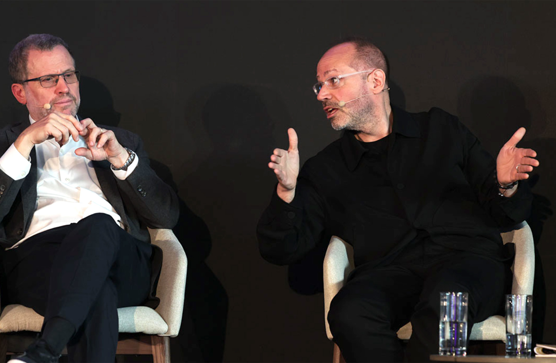 Panel discussion with two speakers engaged in conversation on stage, seated in chairs with microphones.