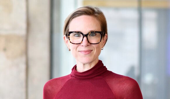 Professional woman with short hair and glasses wearing a maroon turtleneck, smiling in a bright, contemporary setting.